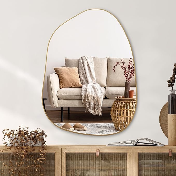 Organic modern asymmetrical wall mirror with slim gold frame, styled above a beige sofa with neutral throw pillows and rattan side table.