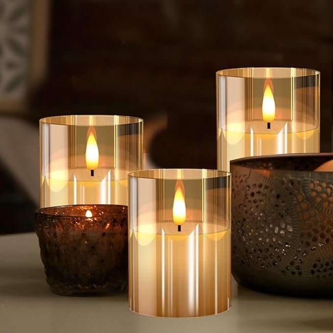 Set of gold glass flameless LED candles with warm flickering light next to decorative metallic and textured candle holders.