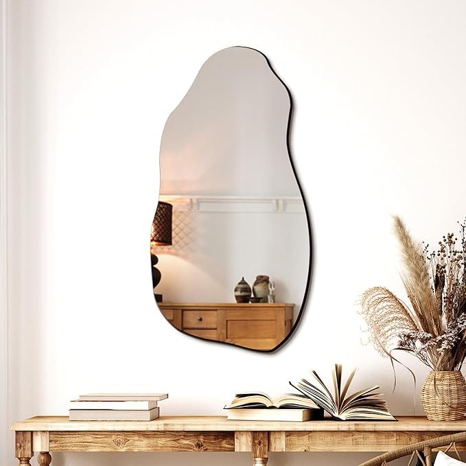 Irregular freeform frameless wall mirror above a rustic wooden console table with dried pampas grass and open books.