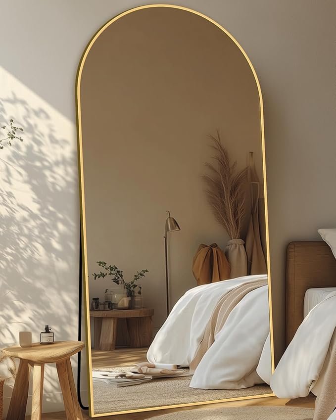 Tall arched floor mirror with thin gold frame leaning against a bedroom wall, reflecting a neutral bed and pampas grass decor.