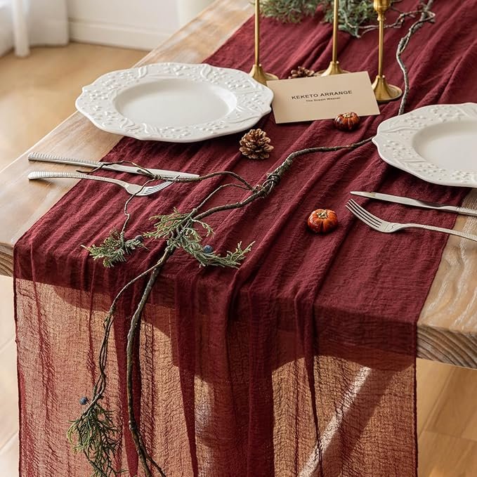 Burgundy sheer table runner styled on a holiday dining table with white plates, pinecones, and greenery accents.