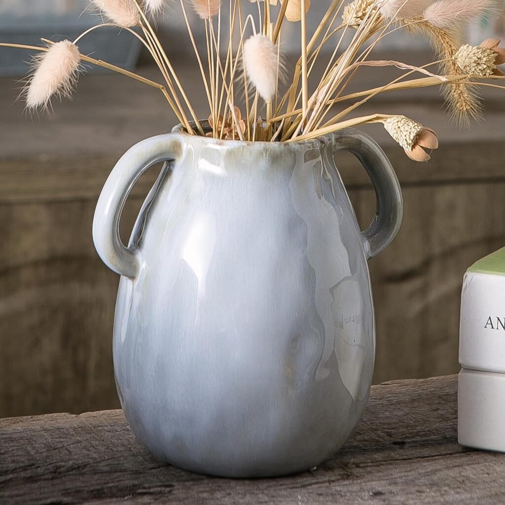 Gray ceramic vase with side handles filled with dried grasses and displayed on a rustic wooden surface.