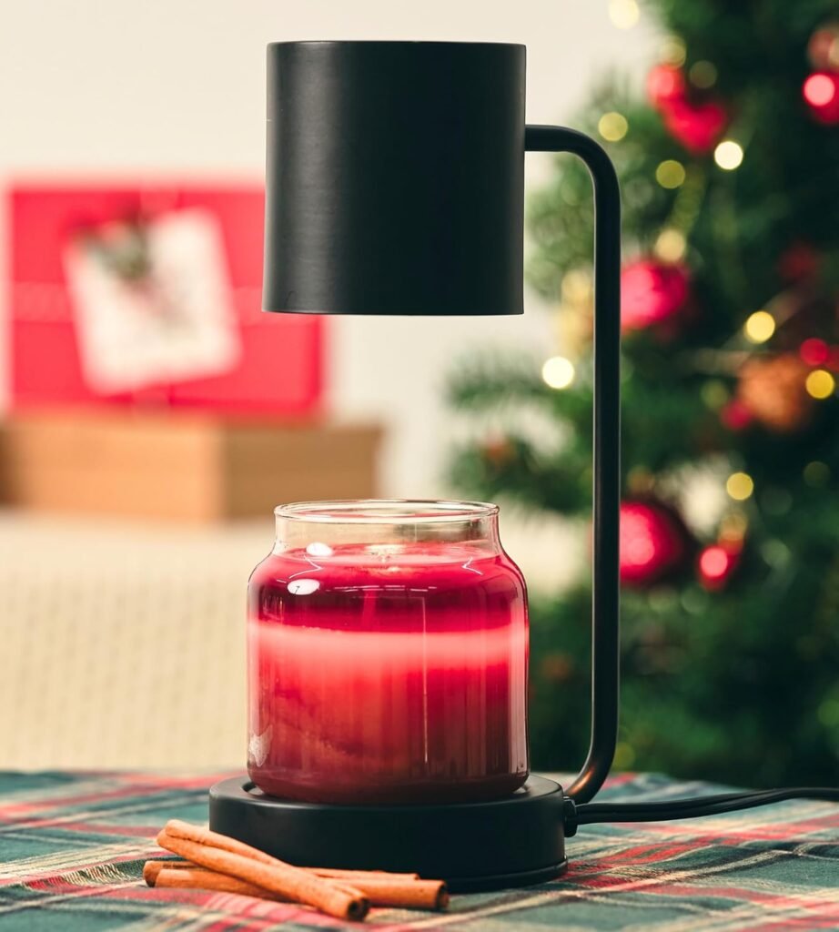Black candle warmer lamp with a red holiday candle glowing beneath, styled with cinnamon sticks and Christmas decor in the background.