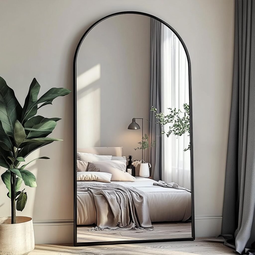 tall arched floor mirror with slim black frame reflecting a neutral bedroom with layered bedding
